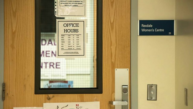 A close-up of a wooden door is seen along with a portion of a sign reading "Rexdale Women's Centre" read on a sign through the window on the door